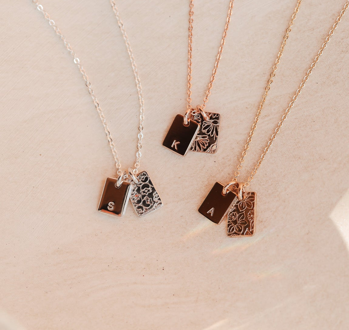 Three necklaces with small rectangular initial pendants on a light pink background