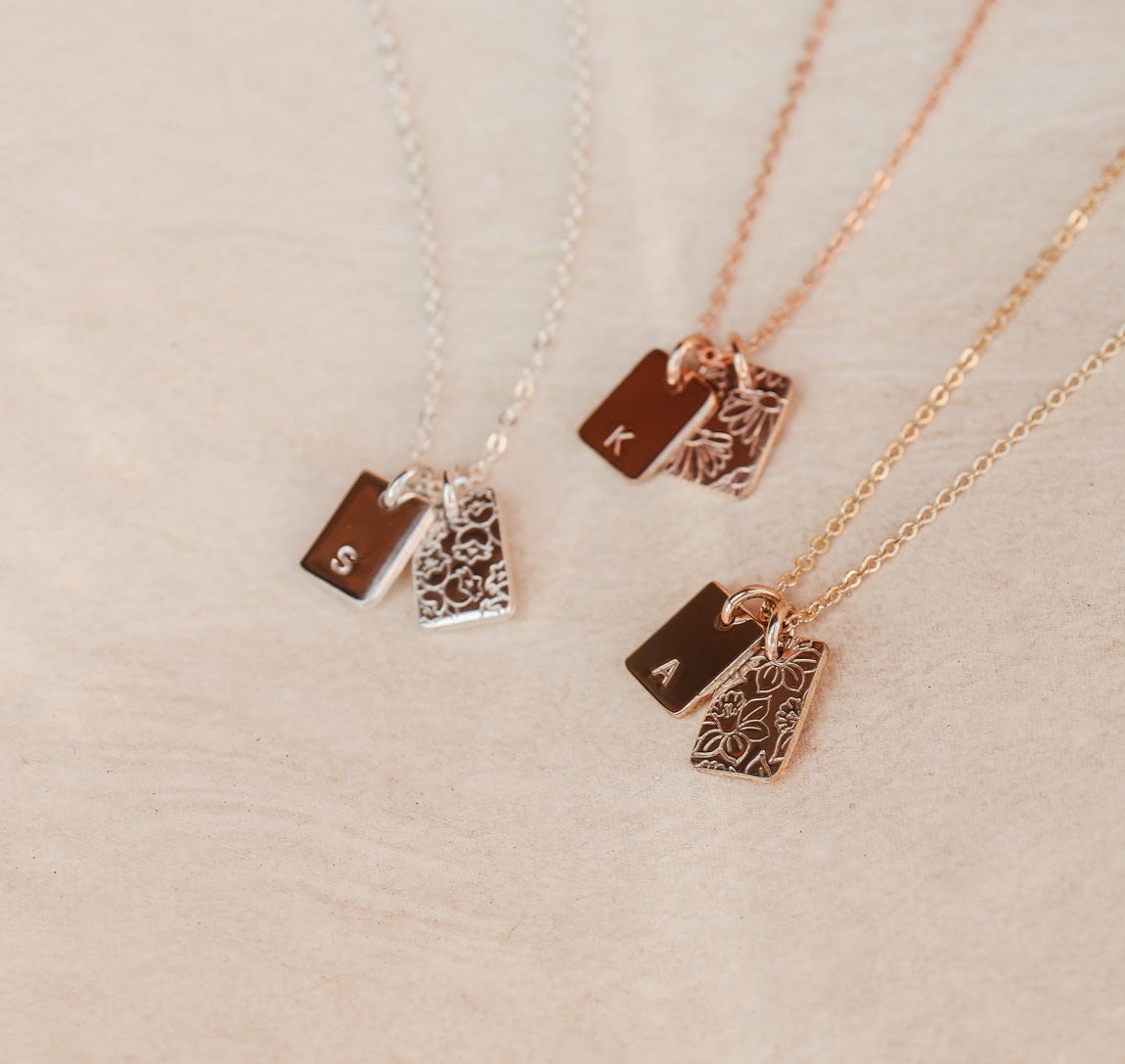 Three gold necklaces with unique initial and birthflower pendants on a textured beige surface